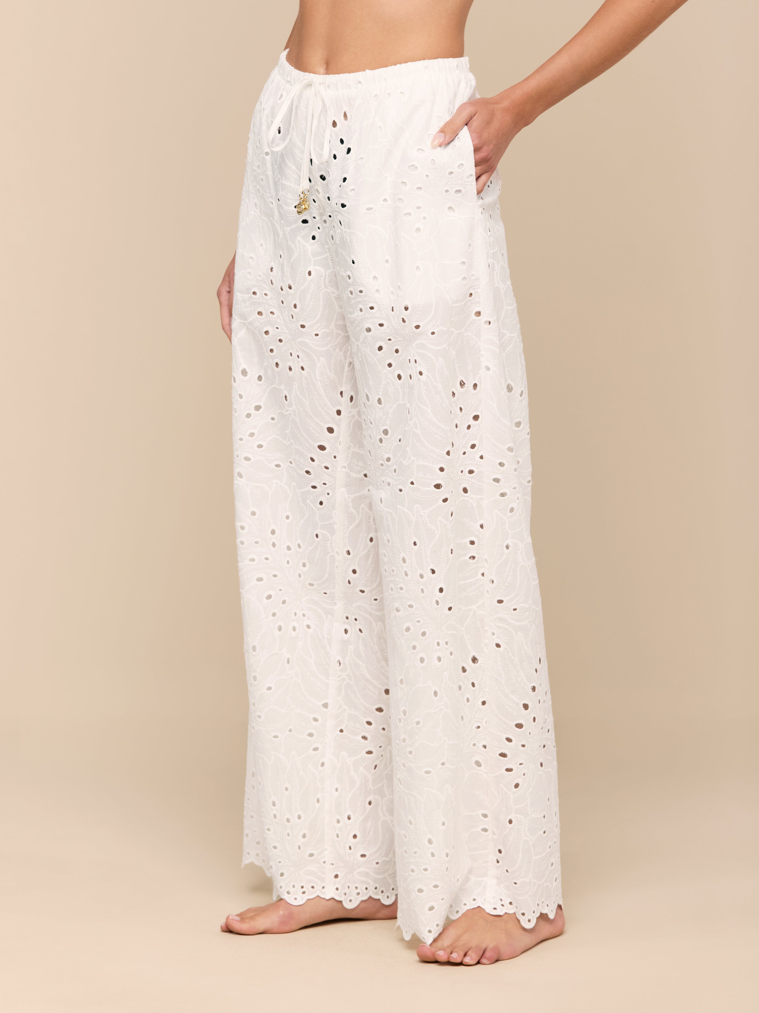 Barefoot, a person models the Geraldine Eyelet Cover-Up Pant—high-waisted, wide-leg cotton pants featuring a tie waist. The image focuses on the lower torso and legs set against a plain, light background.