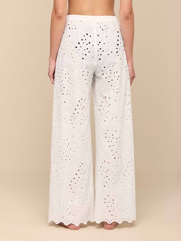 A person stands barefoot, facing away, wearing the Geraldine Eyelet Cover-Up Pant—high-waisted, white cotton eyelet pants featuring a scalloped hem. Elegant and designed for resort wear, these semi-sheer pants showcase a delicate floral cutout pattern.