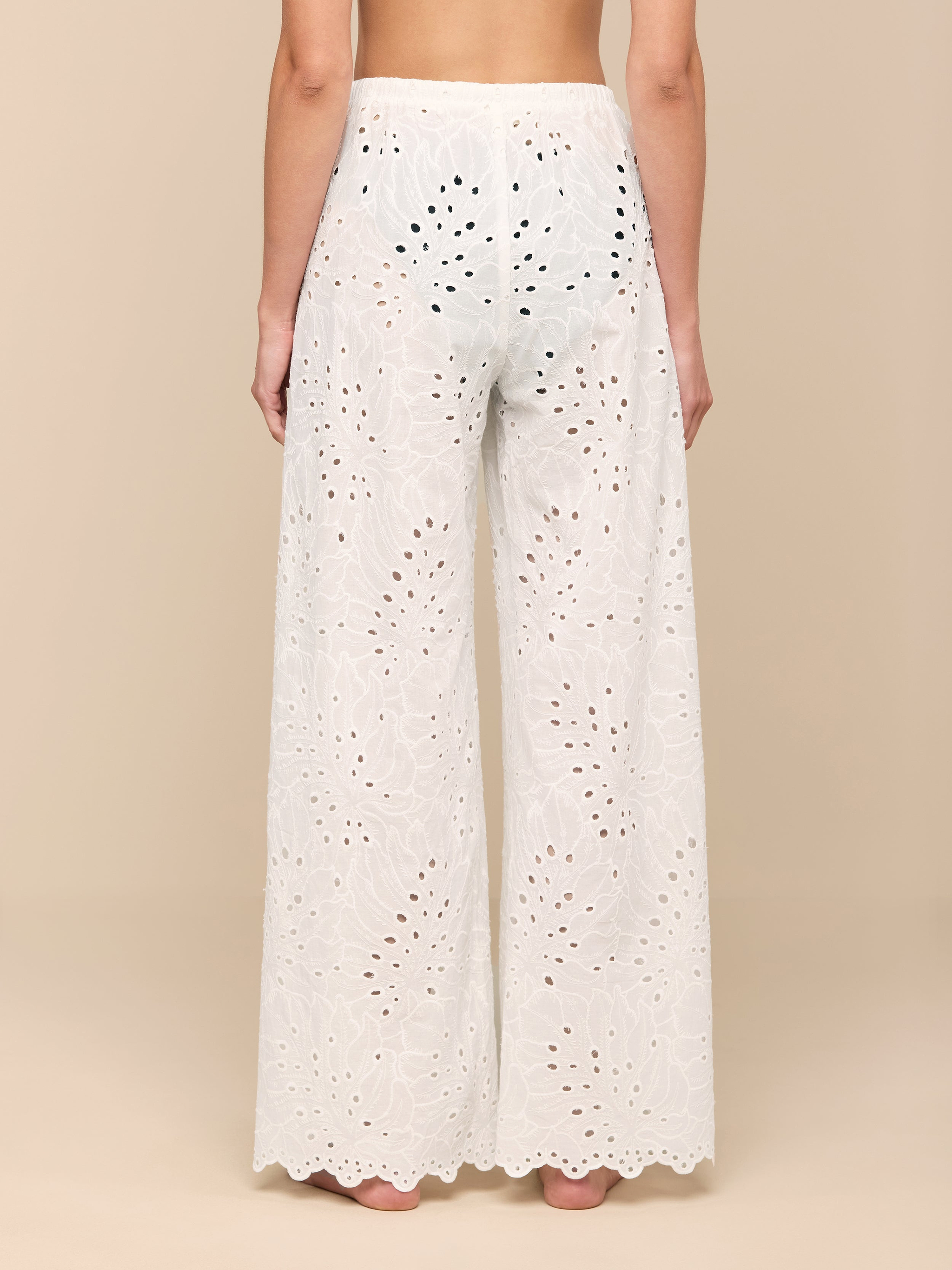 A person stands barefoot, facing away, wearing the Geraldine Eyelet Cover-Up Pant—high-waisted, white cotton eyelet pants featuring a scalloped hem. Elegant and designed for resort wear, these semi-sheer pants showcase a delicate floral cutout pattern.
