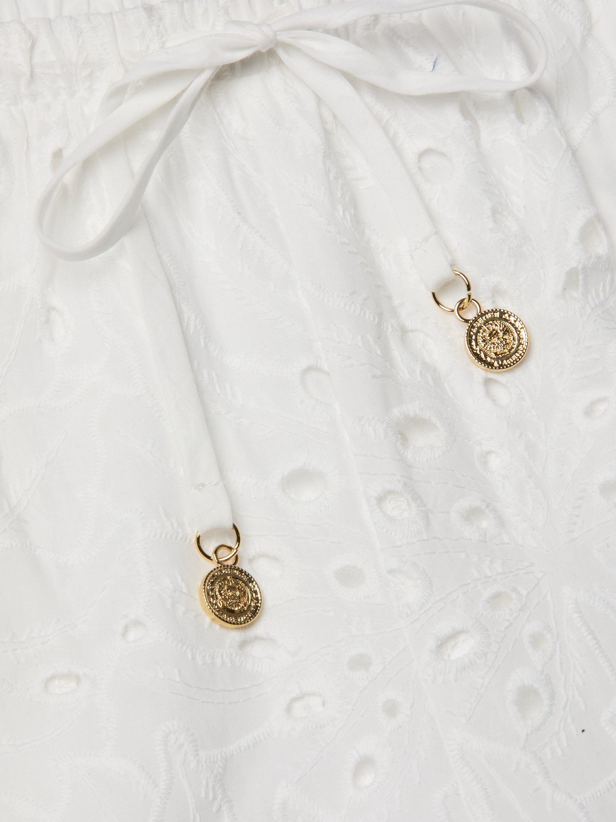 Close-up of the Geraldine Eyelet Cover-Up Pant, crafted from white cotton eyelet fabric with a tie waist and drawstring bow. Two gold decorative pendants accent the drawstring ends, bringing an elegant touch to your resort wear ensemble.