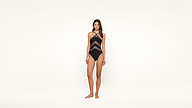 A woman stands barefoot on a white background wearing a black one-piece swimsuit with sheer diagonal stripes and crisscross straps.
