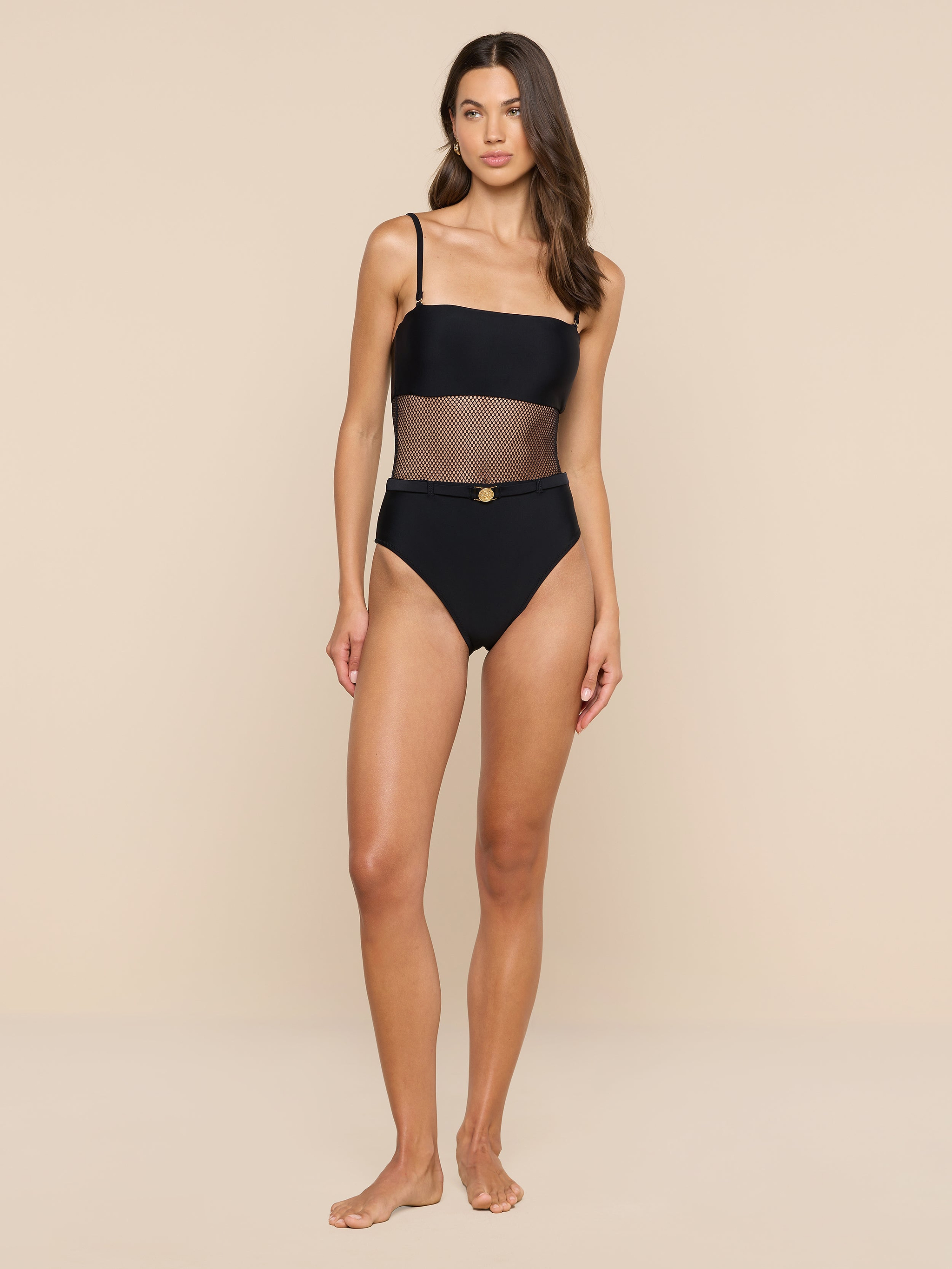 A woman stands barefoot against a white background, wearing the Tory Bandeau One-Piece Swimsuit, which features a one-shoulder design with mesh insets at the waist and high-waisted bottoms.