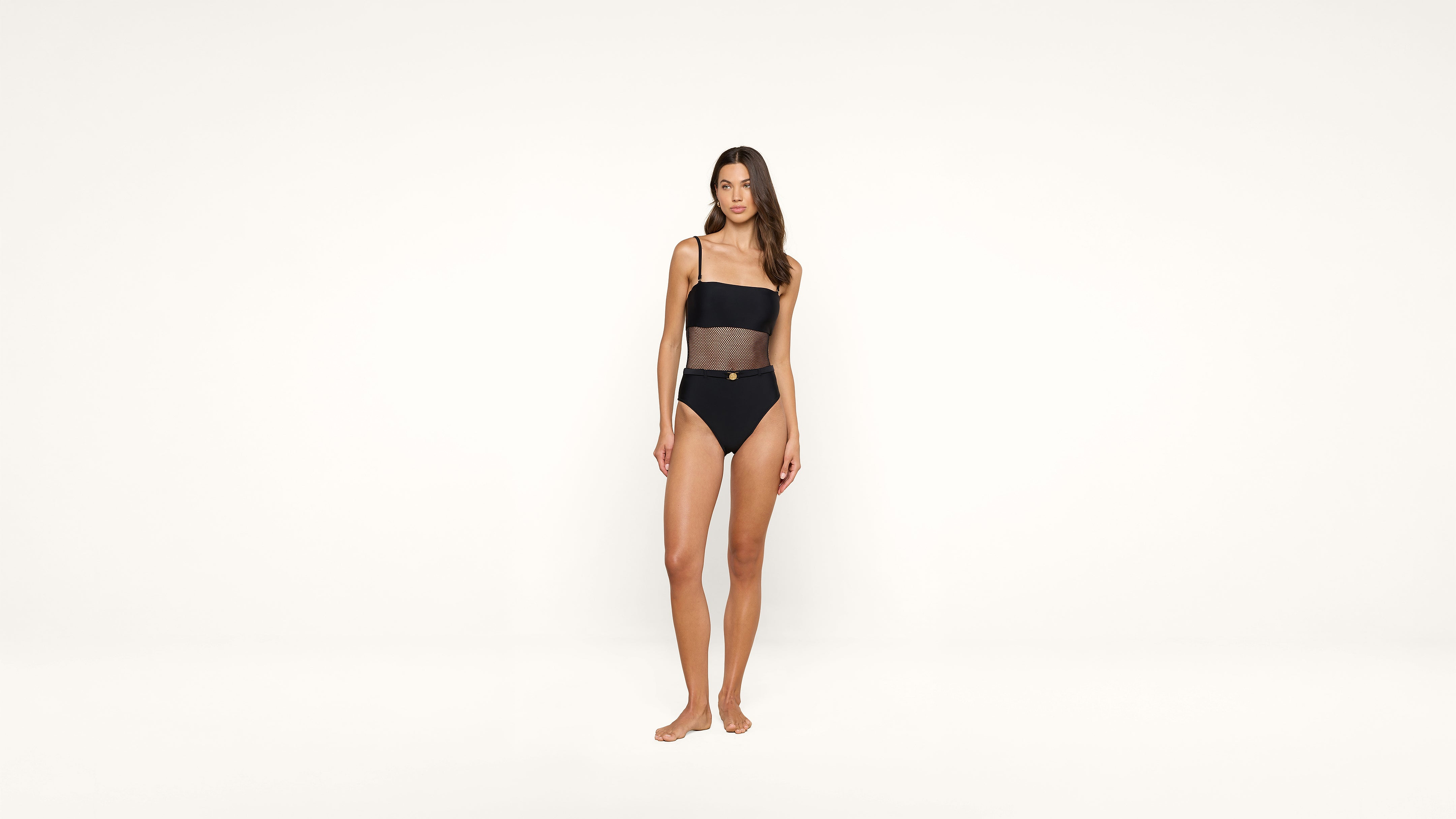 A woman stands barefoot against a plain white background, wearing a black one-piece swimsuit with sheer panel detailing and thin shoulder straps.