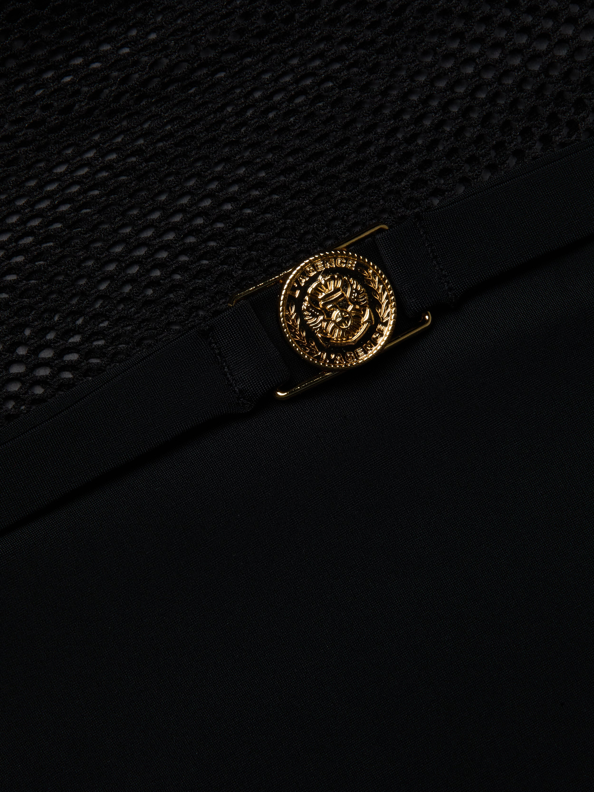 A close-up of a gold Medusa head emblem on the black fabric of the Tory Bandeau One-Piece Swimsuit, shown above a section of black mesh insets.