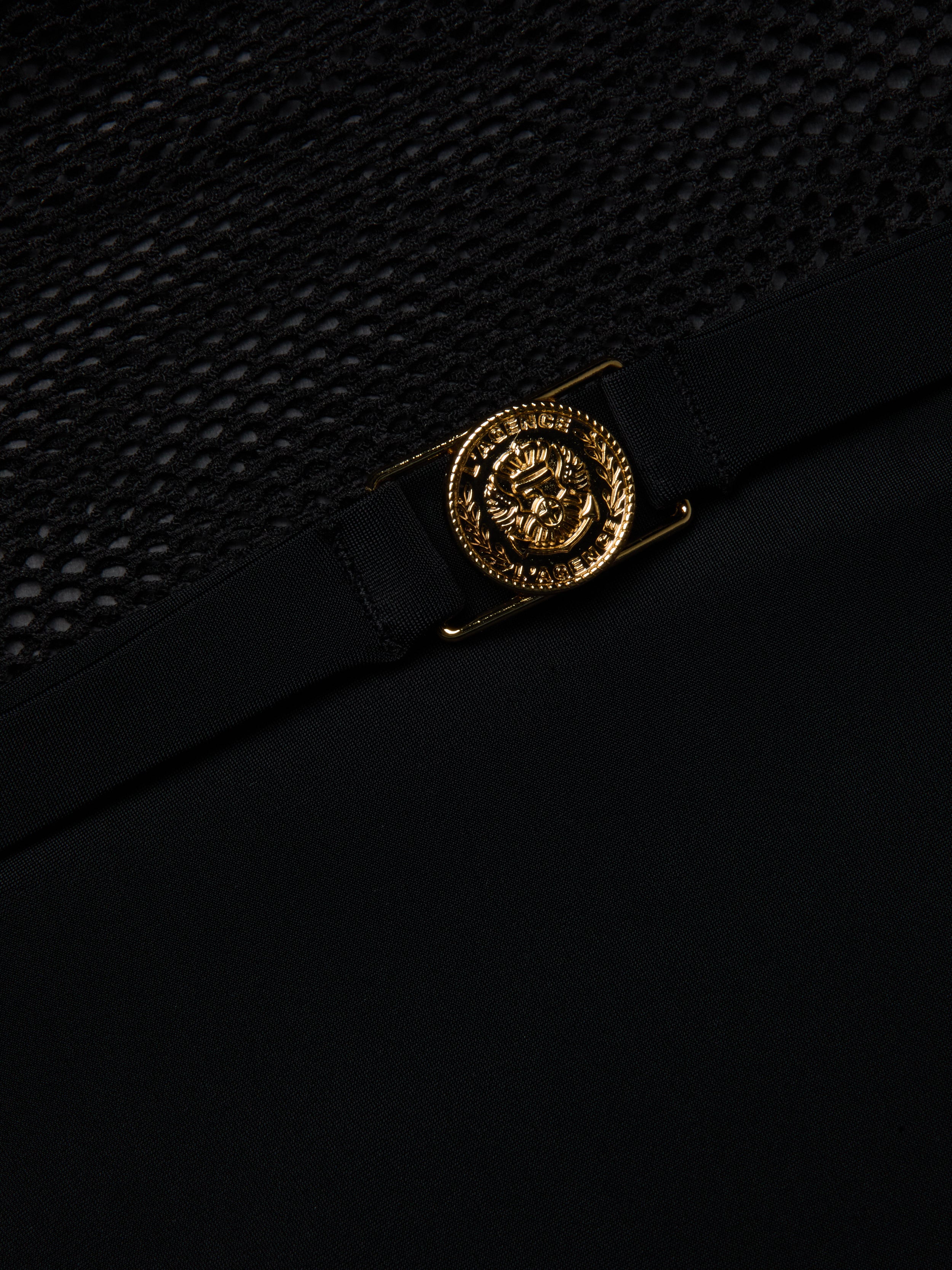 A close-up of a gold Medusa head emblem on the black fabric of the Tory Bandeau One-Piece Swimsuit, shown above a section of black mesh insets.