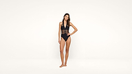 A woman stands against a plain white background wearing a black one-piece swimsuit with sheer mesh panels and a halter neck design. One hand is on her hip, and she looks confidently at the camera.