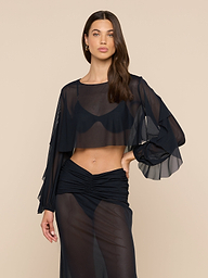 A woman with long brown hair wears the Ambala Mesh Cover-Up Top, a sheer black cropped silhouette with long, loose sleeves, paired with a matching sheer skirt over a black bra and underwear against a plain beige background.