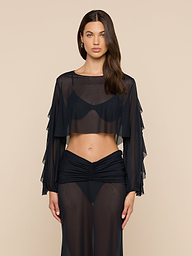 A woman stands against a beige background wearing the Ambala Mesh Cover-Up Top, a sheer black long-sleeve crop top with ruffle details, paired with matching high-waisted sheer pants for a chic, modern look.