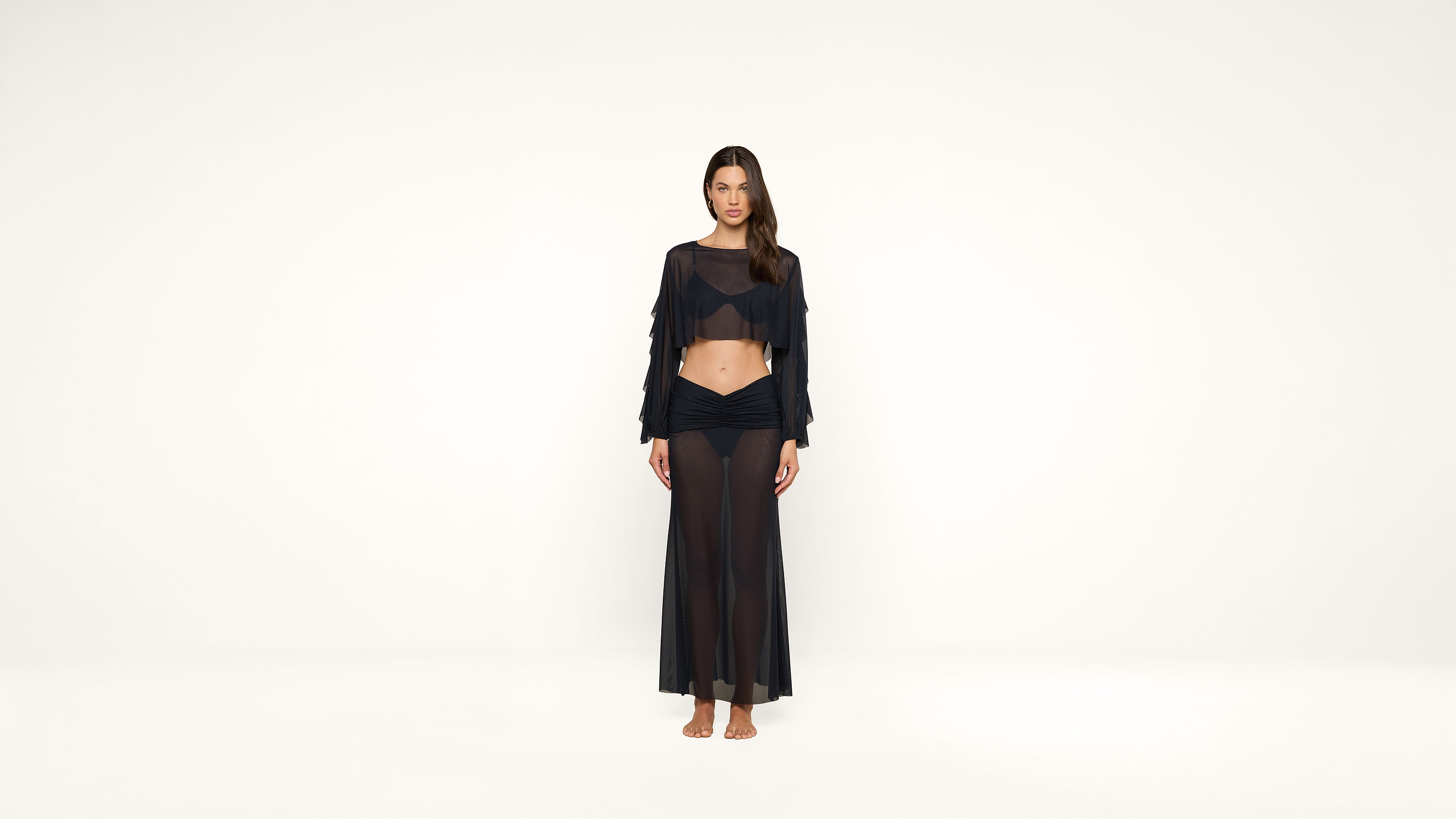 A woman stands barefoot against a white background, wearing a sheer black long-sleeve crop top and a matching sheer black maxi skirt.