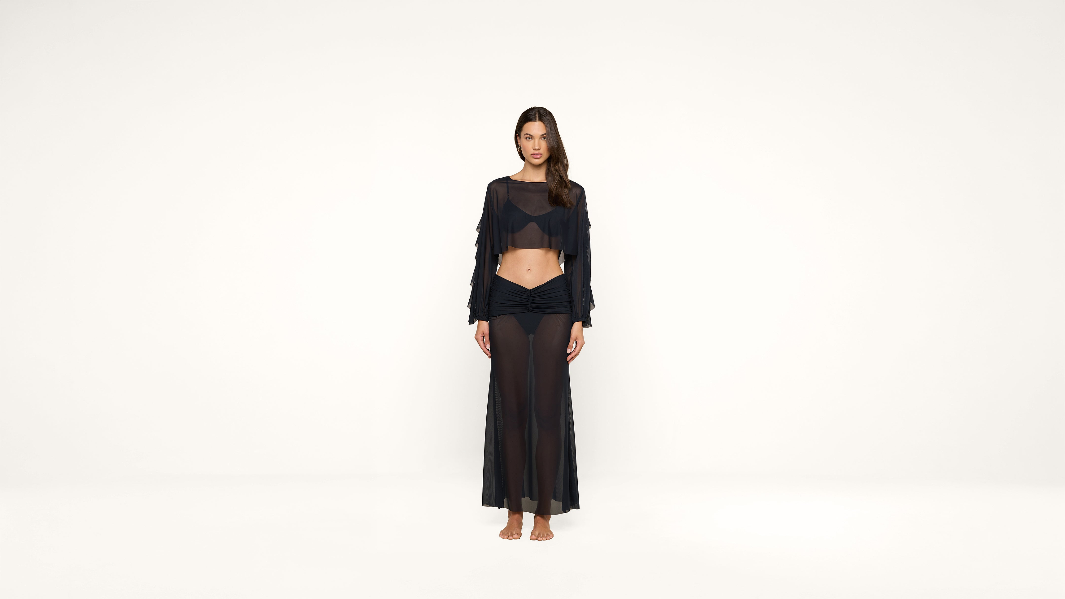 A woman stands barefoot against a white background, wearing a sheer black long-sleeve crop top and a matching sheer black maxi skirt.