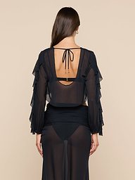 A woman with long brown hair is seen from the back wearing the Ambala Mesh Cover-Up Top, a sheer black piece with ruffled long sleeves and a tied open back, revealing her black bra and high-waisted underwear underneath.