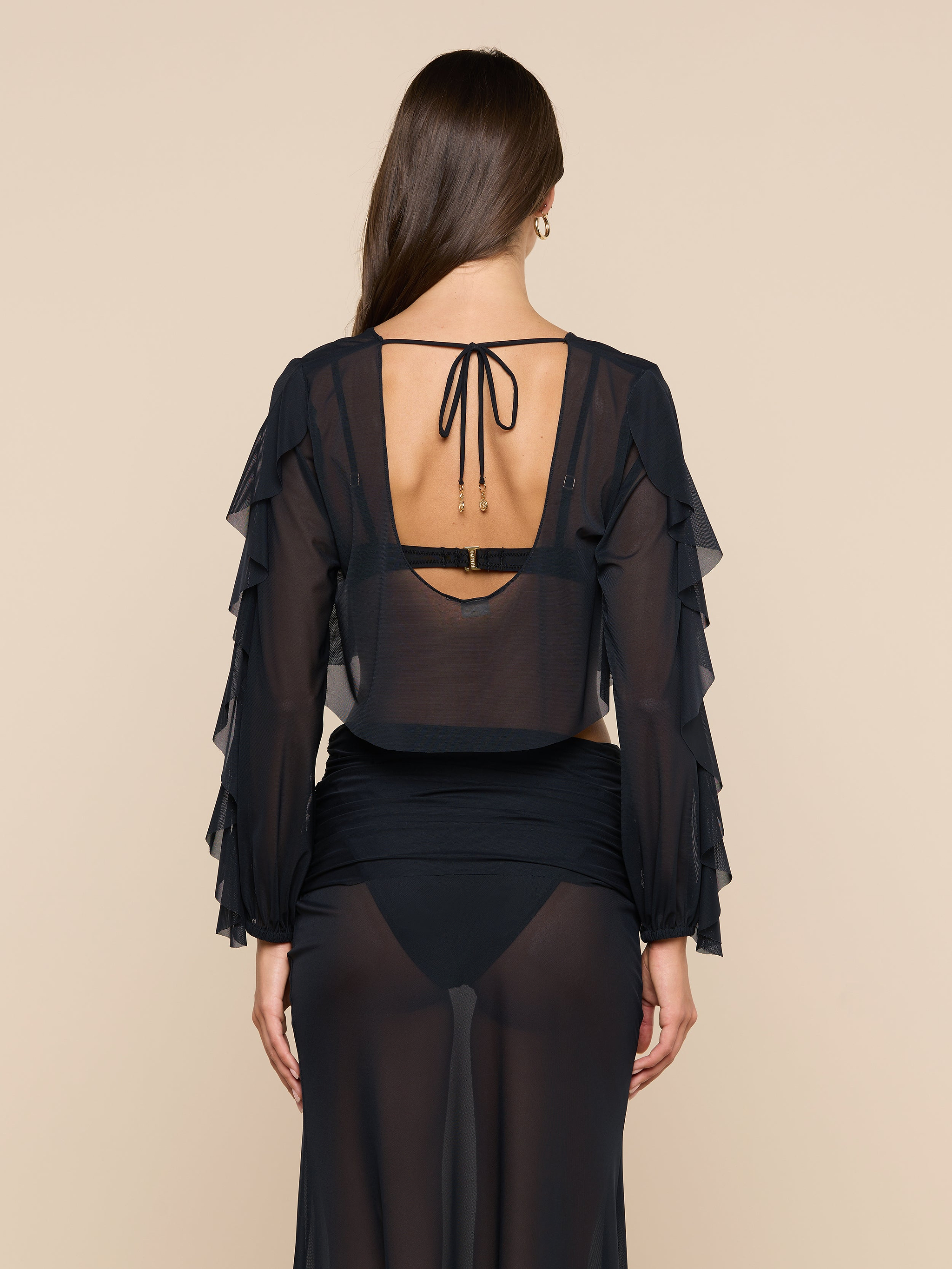 A woman with long brown hair is seen from the back wearing the Ambala Mesh Cover-Up Top, a sheer black piece with ruffled long sleeves and a tied open back, revealing her black bra and high-waisted underwear underneath.
