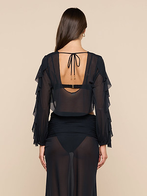 A woman with long brown hair is seen from the back wearing the Ambala Mesh Cover-Up Top, a sheer black piece with ruffled long sleeves and a tied open back, revealing her black bra and high-waisted underwear underneath.