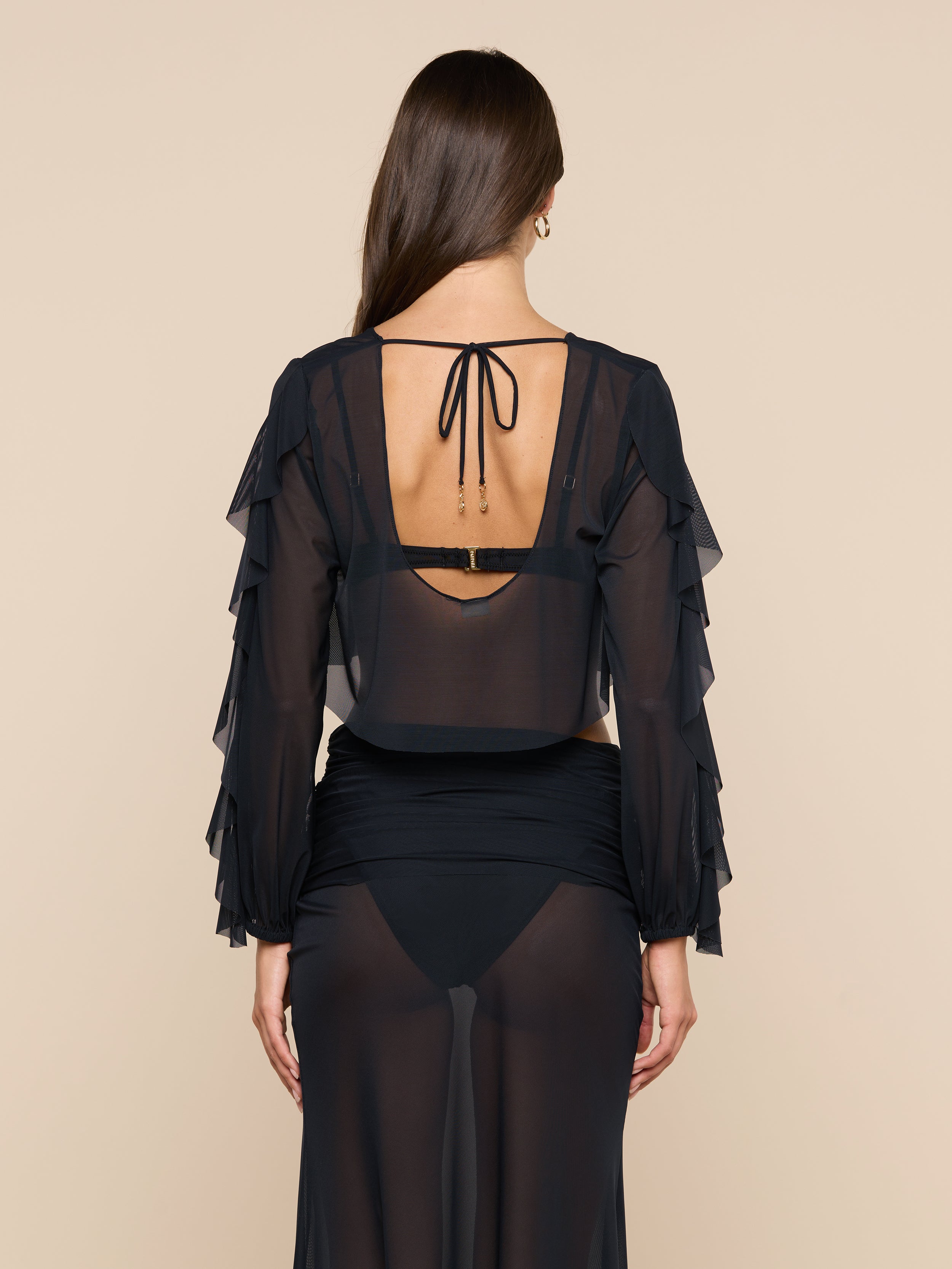 A woman with long brown hair is seen from the back wearing the Ambala Mesh Cover-Up Top, a sheer black piece with ruffled long sleeves and a tied open back, revealing her black bra and high-waisted underwear underneath.