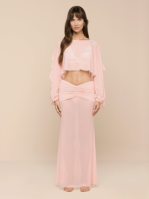 A woman stands barefoot against a beige background, wearing the light pink Ambala Cover-Up Top—a sheer nylon, long-sleeve crop with ruffles—embodying effortless beachside style as she faces the camera.