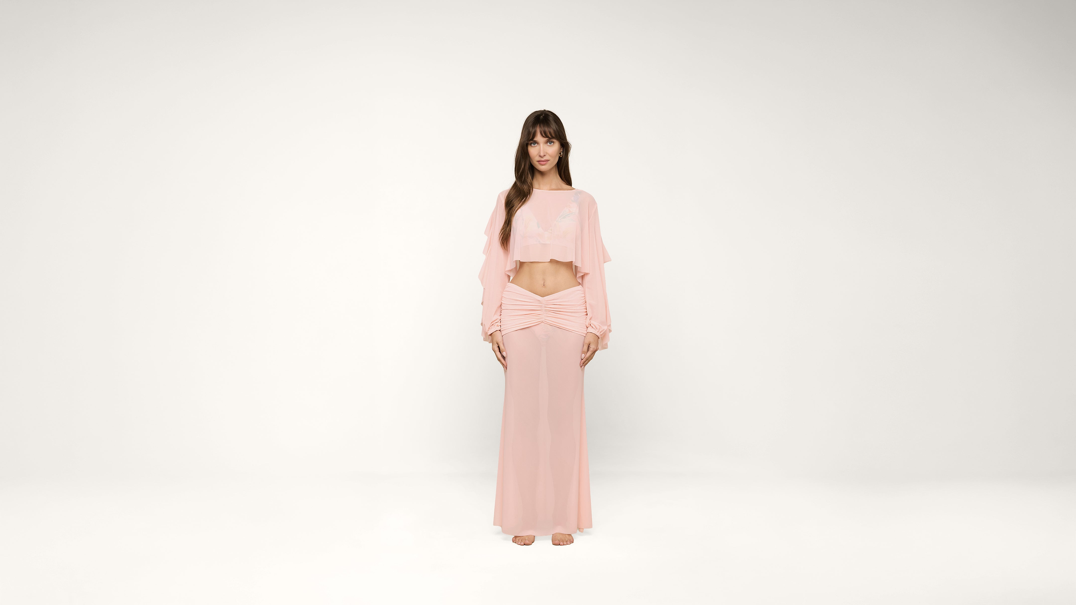 A woman stands barefoot against a white background, wearing a light pink, long-sleeved crop top and a matching long skirt. She has long, brown hair with bangs and a neutral expression.