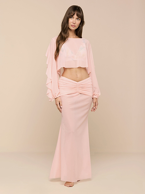A woman stands barefoot in a sheer, ruffled, long-sleeve Ambala Cover-Up Top and flowing light pink nylon skirt, capturing effortless beachside style against a plain beige background.