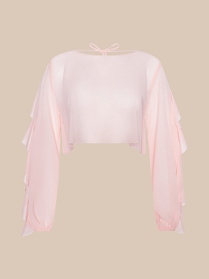 The Ambala Cover-Up Top is a sheer light pink long-sleeve crop top in delicate nylon with ruffled sleeves, round neckline, and back neck tie—ideal for effortless beachside style. Shown on a beige background.