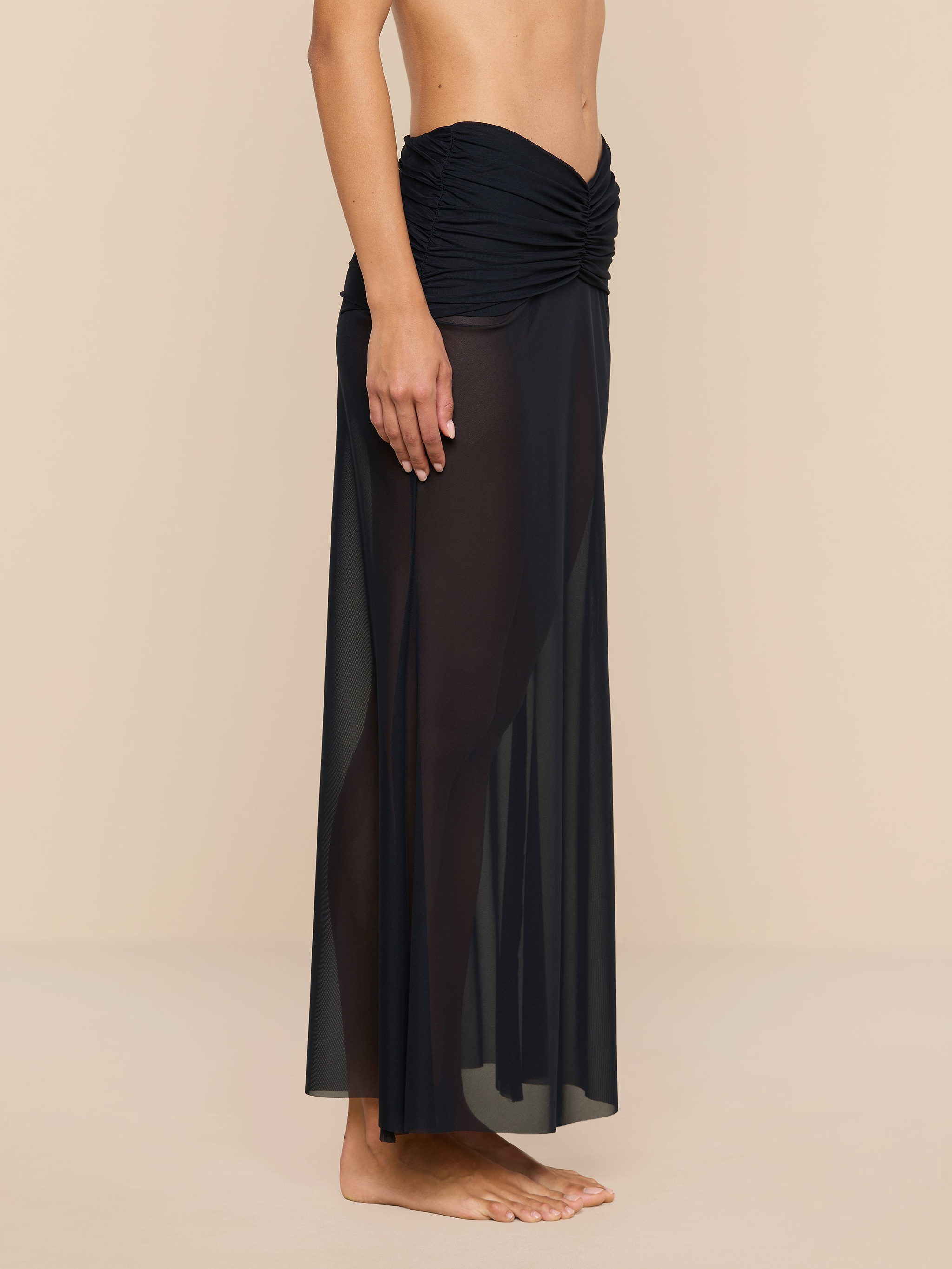 A person stands barefoot, shown from the chest down, wearing the Mahe Mesh Cover-Up Skirt—a black, sheer mermaid skirt with a ruched waistband—perfect as a chic poolside layer. The background is plain beige.