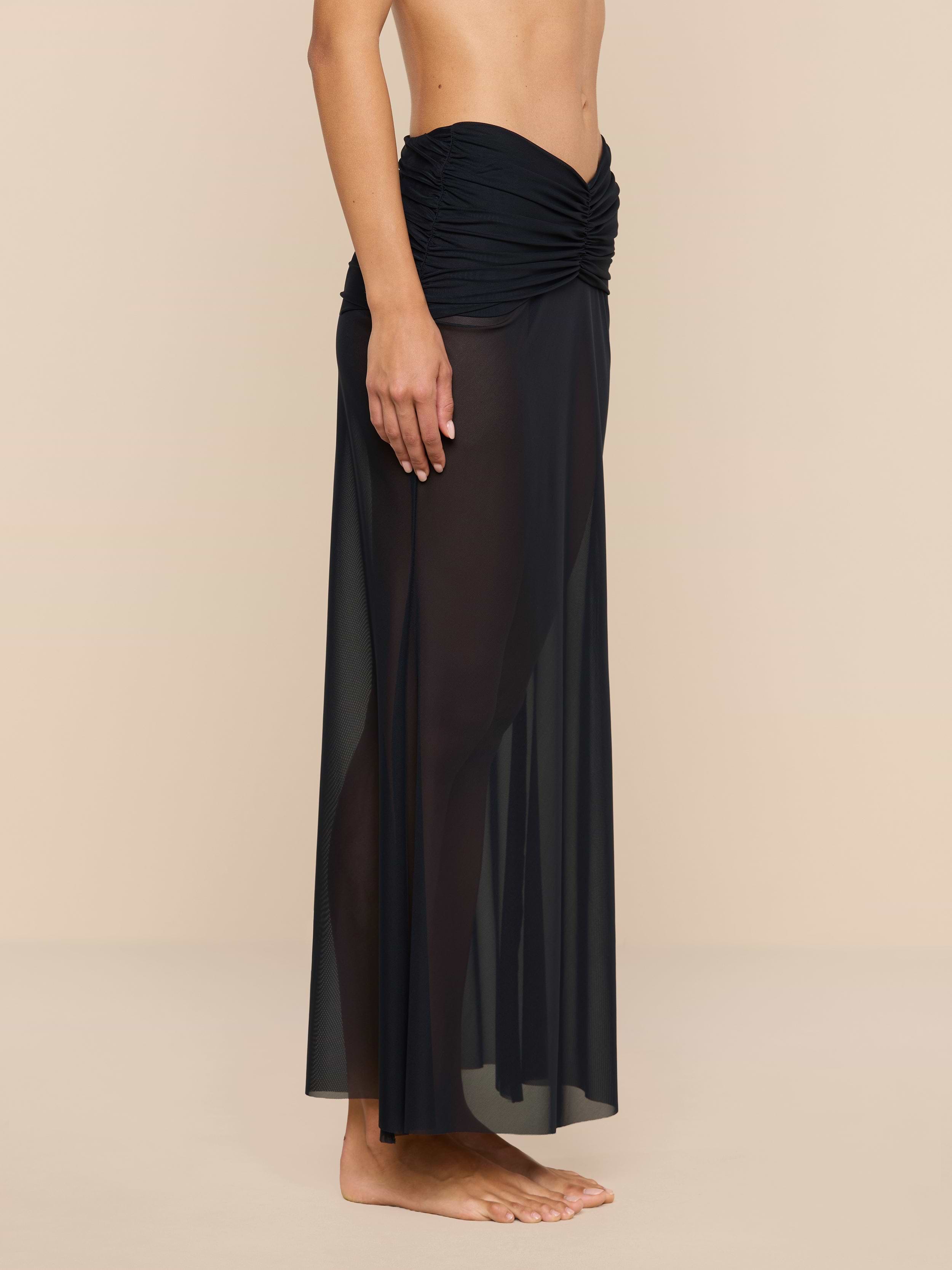 A person stands barefoot, shown from the chest down, wearing the Mahe Mesh Cover-Up Skirt—a black, sheer mermaid skirt with a ruched waistband—perfect as a chic poolside layer. The background is plain beige.