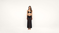 A woman stands barefoot against a plain white background, wearing a black bikini top and a long, sheer black skirt. She faces forward with a neutral expression and her arms relaxed by her sides.