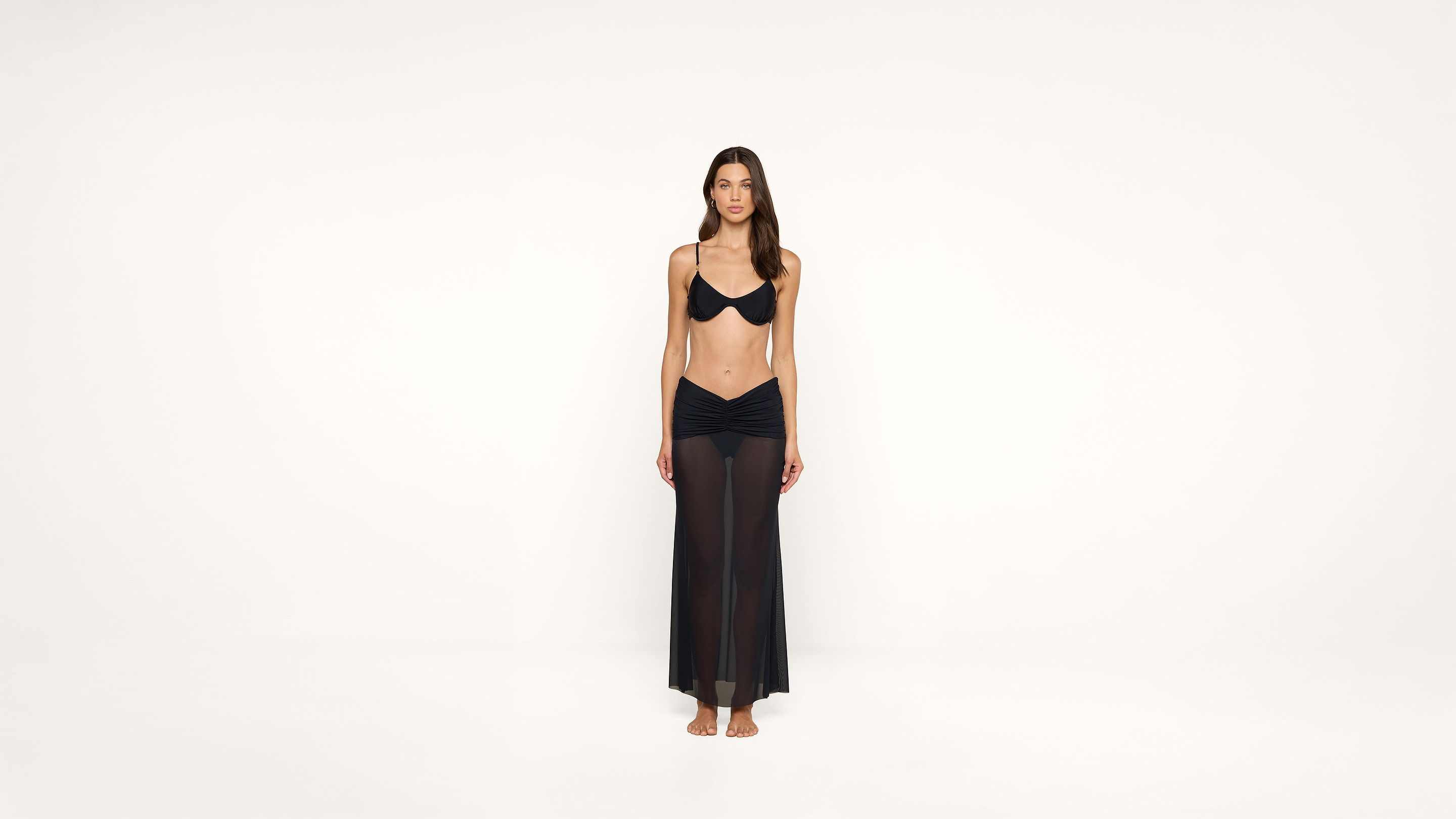 A woman stands barefoot against a plain white background, wearing a black bikini top and a long, sheer black skirt. She faces forward with a neutral expression and her arms relaxed by her sides.