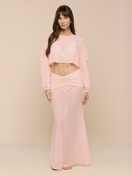 A woman stands in a light pink, long-sleeve crop top and the Mahe Mesh Cover-Up Skirt, styled as a matching set that channels poolside elegance. She faces forward with a neutral expression against a plain white background.