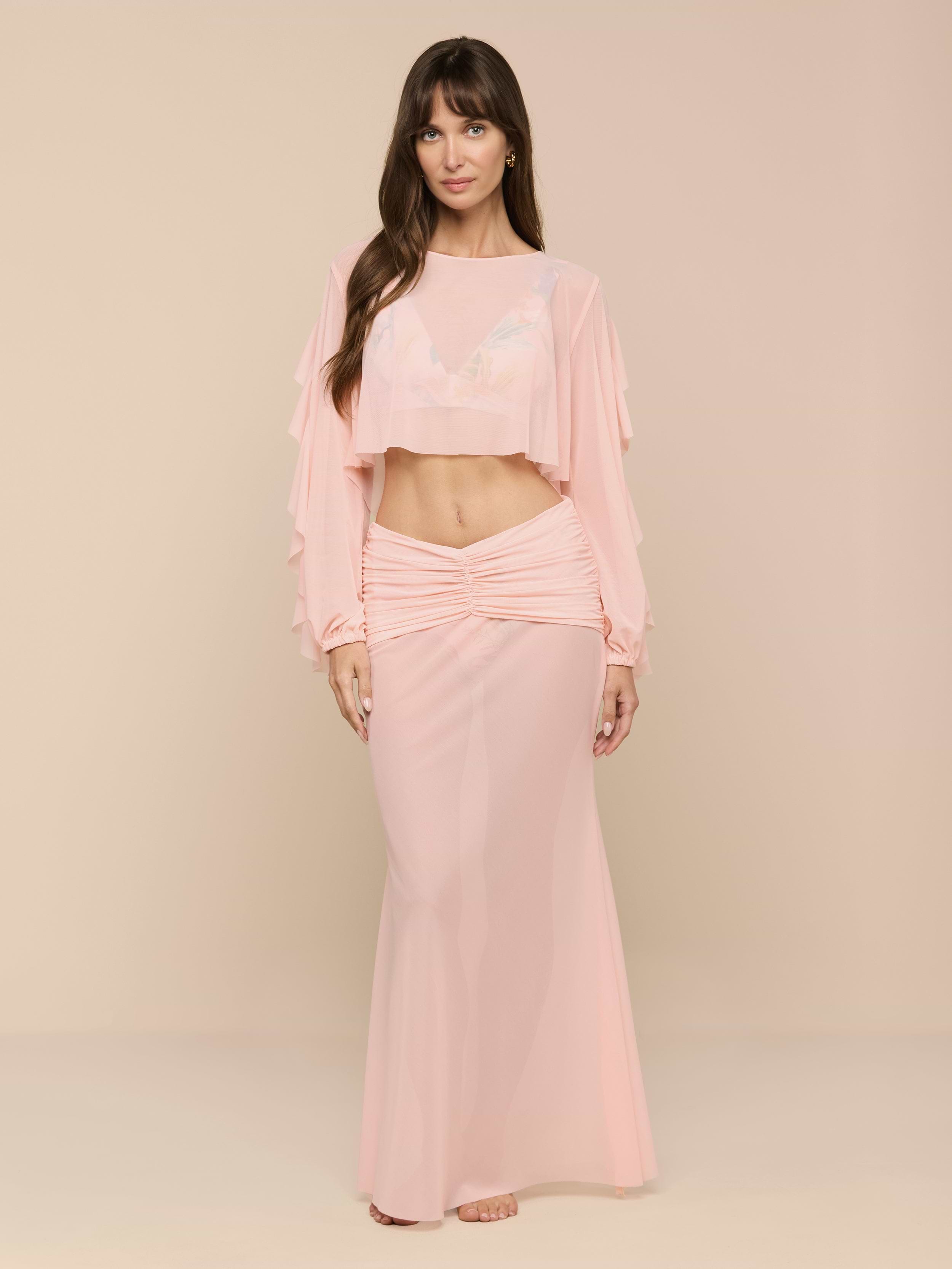 A woman stands in a light pink, long-sleeve crop top and the Mahe Mesh Cover-Up Skirt, styled as a matching set that channels poolside elegance. She faces forward with a neutral expression against a plain white background.