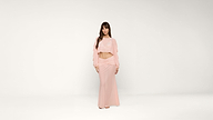 A woman stands against a plain white background, wearing a light pink long-sleeve crop top and matching floor-length skirt. She has long brown hair and is looking at the camera with a neutral expression.