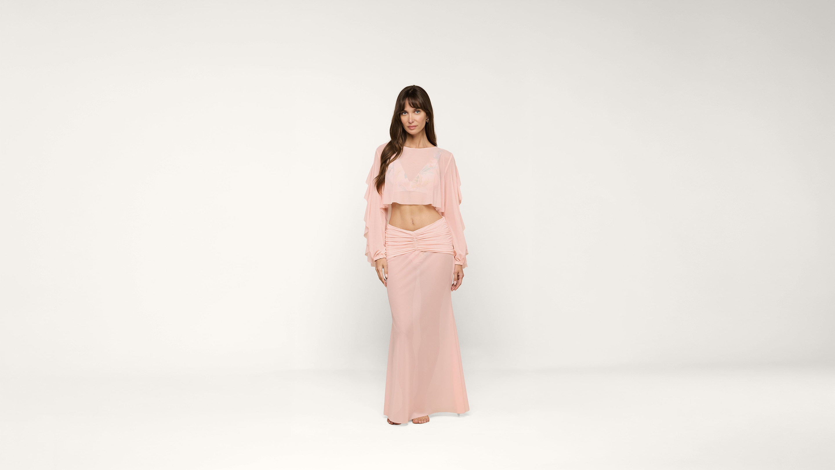 A woman stands against a plain white background, wearing a light pink long-sleeve crop top and matching floor-length skirt. She has long brown hair and is looking at the camera with a neutral expression.