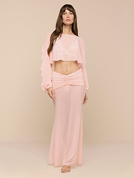 A woman stands barefoot in a minimalist space, wearing the Mahe Mesh Cover-Up Skirt—a flowing, light pink, high-waisted mermaid skirt—paired with a matching long-sleeve crop top with ruffled sleeves.