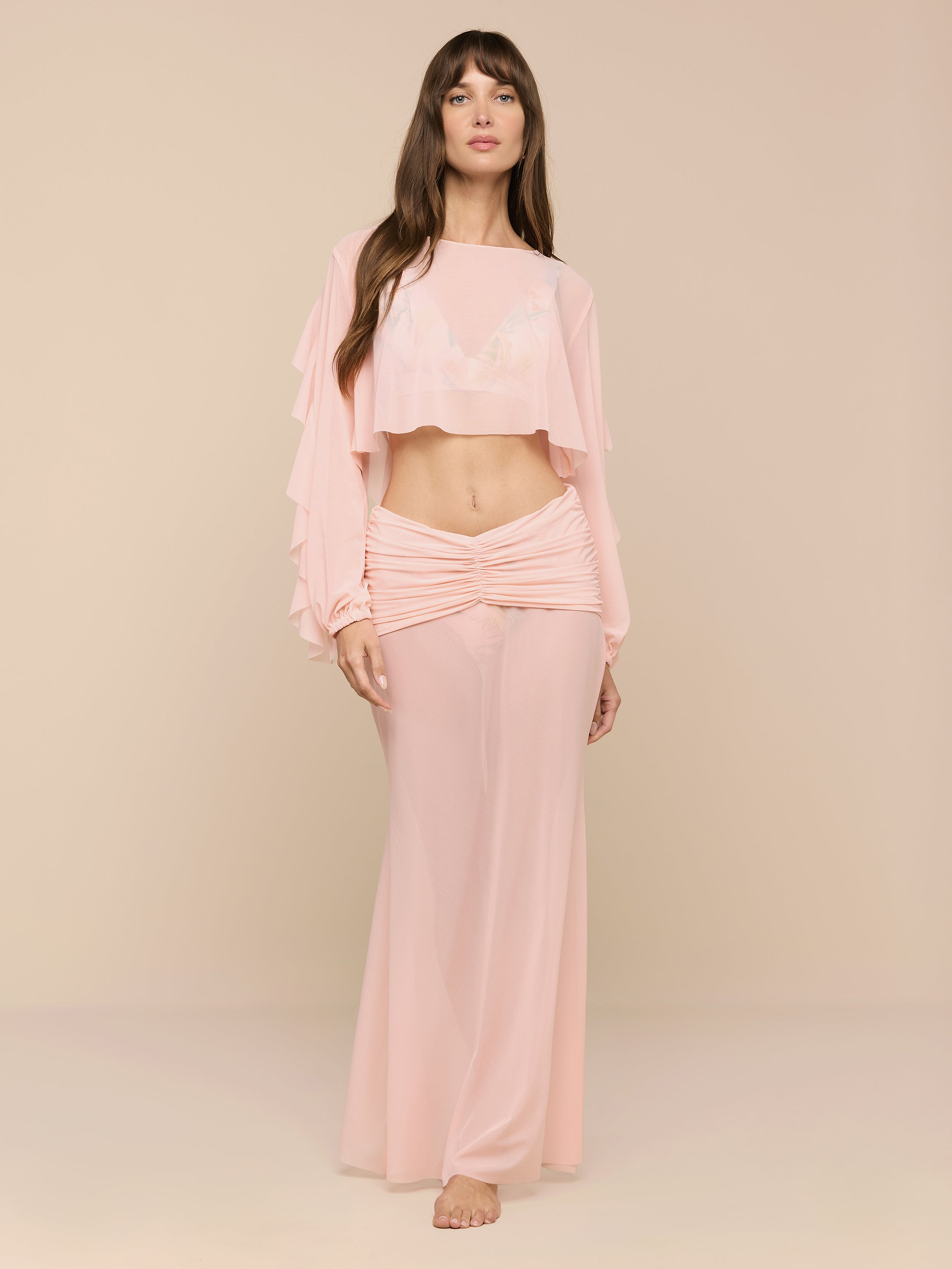 A woman stands barefoot in a minimalist space, wearing the Mahe Mesh Cover-Up Skirt—a flowing, light pink, high-waisted mermaid skirt—paired with a matching long-sleeve crop top with ruffled sleeves.