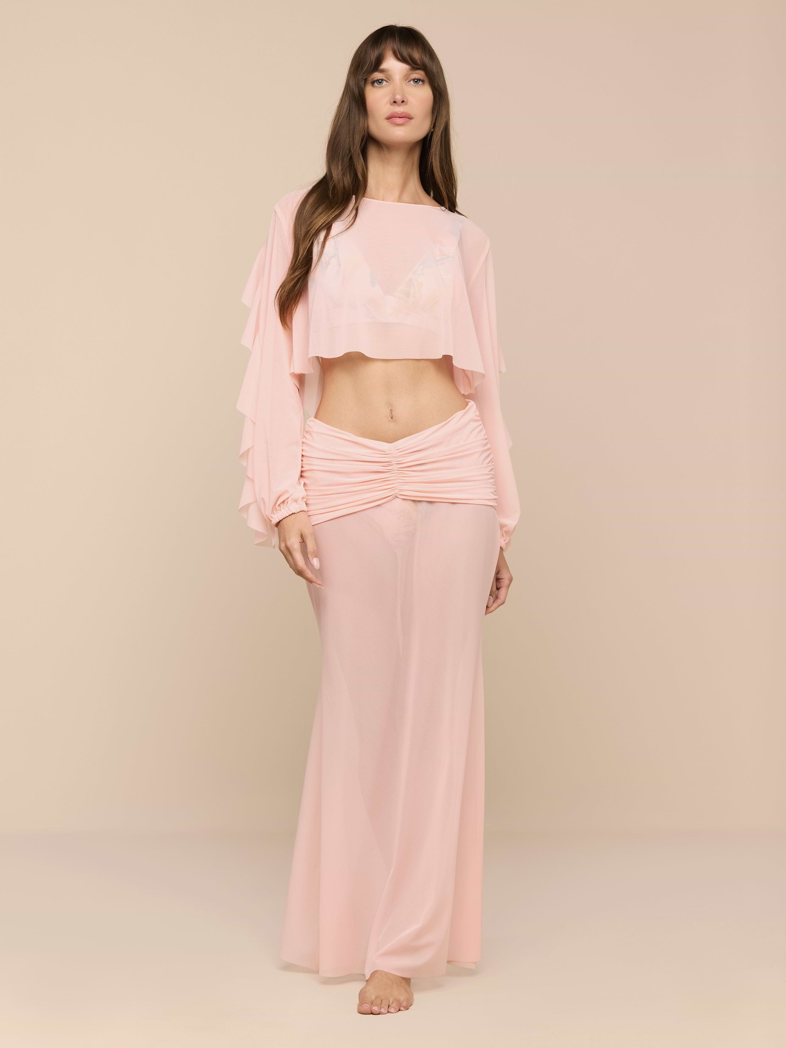 A woman stands barefoot in a minimalist space, wearing the Mahe Mesh Cover-Up Skirt—a flowing, light pink, high-waisted mermaid skirt—paired with a matching long-sleeve crop top with ruffled sleeves.