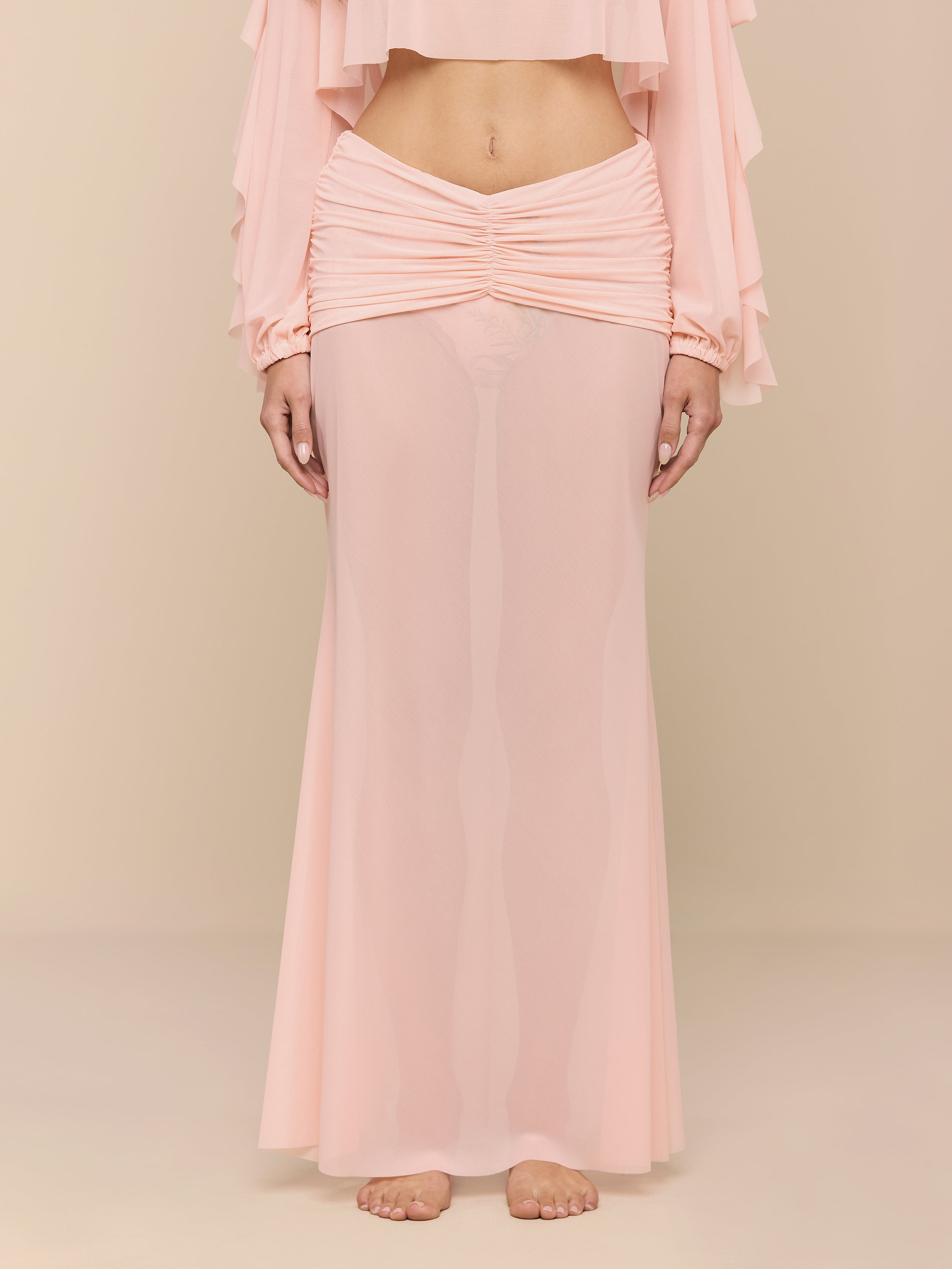 A person stands barefoot on a white background, wearing the light pink Mahe Mesh Cover-Up Skirt styled as a mermaid skirt and a matching long-sleeve top that reveals their midriff. The person’s head is not visible.