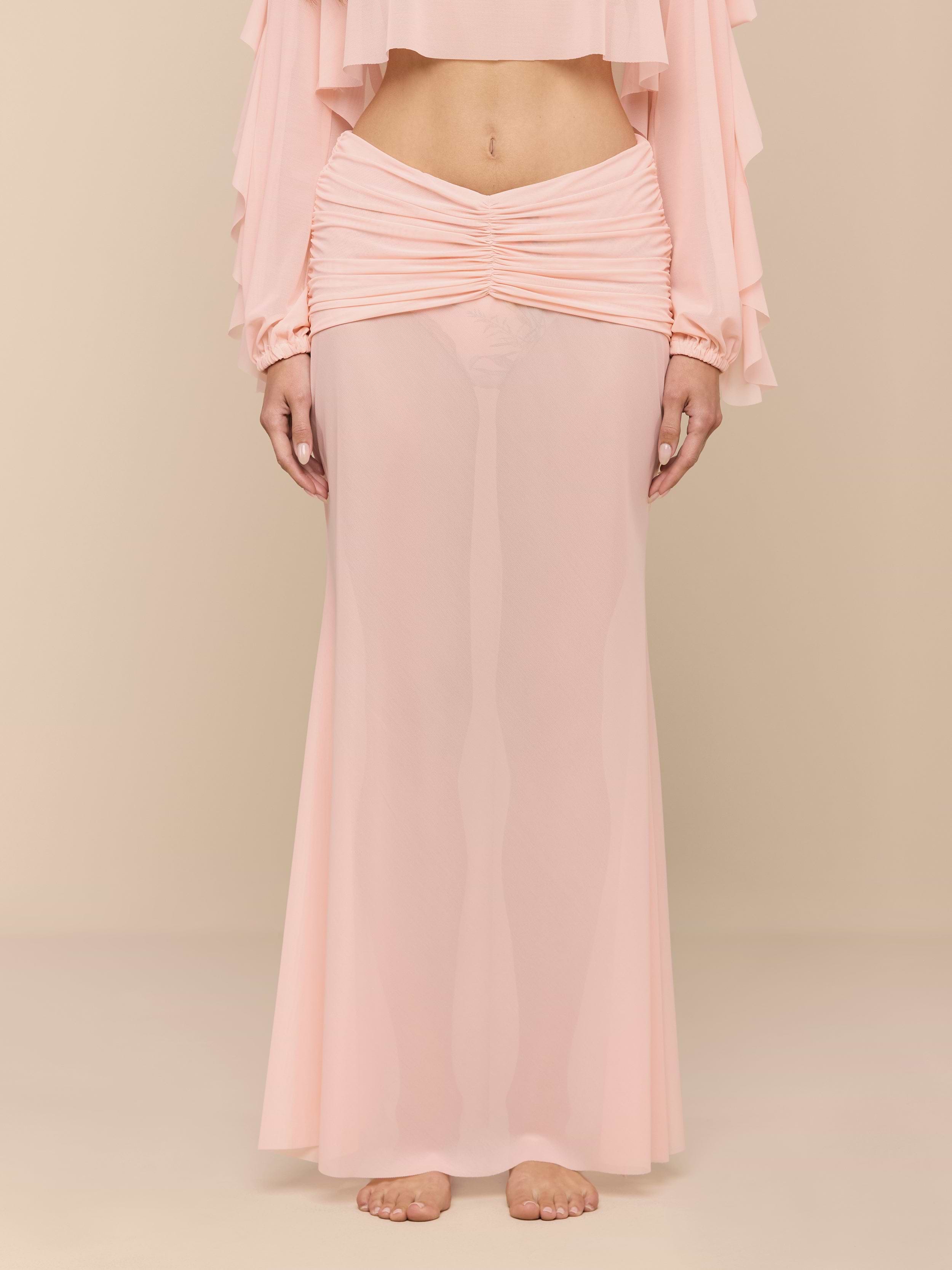 A person stands barefoot on a white background, wearing the light pink Mahe Mesh Cover-Up Skirt styled as a mermaid skirt and a matching long-sleeve top that reveals their midriff. The person’s head is not visible.