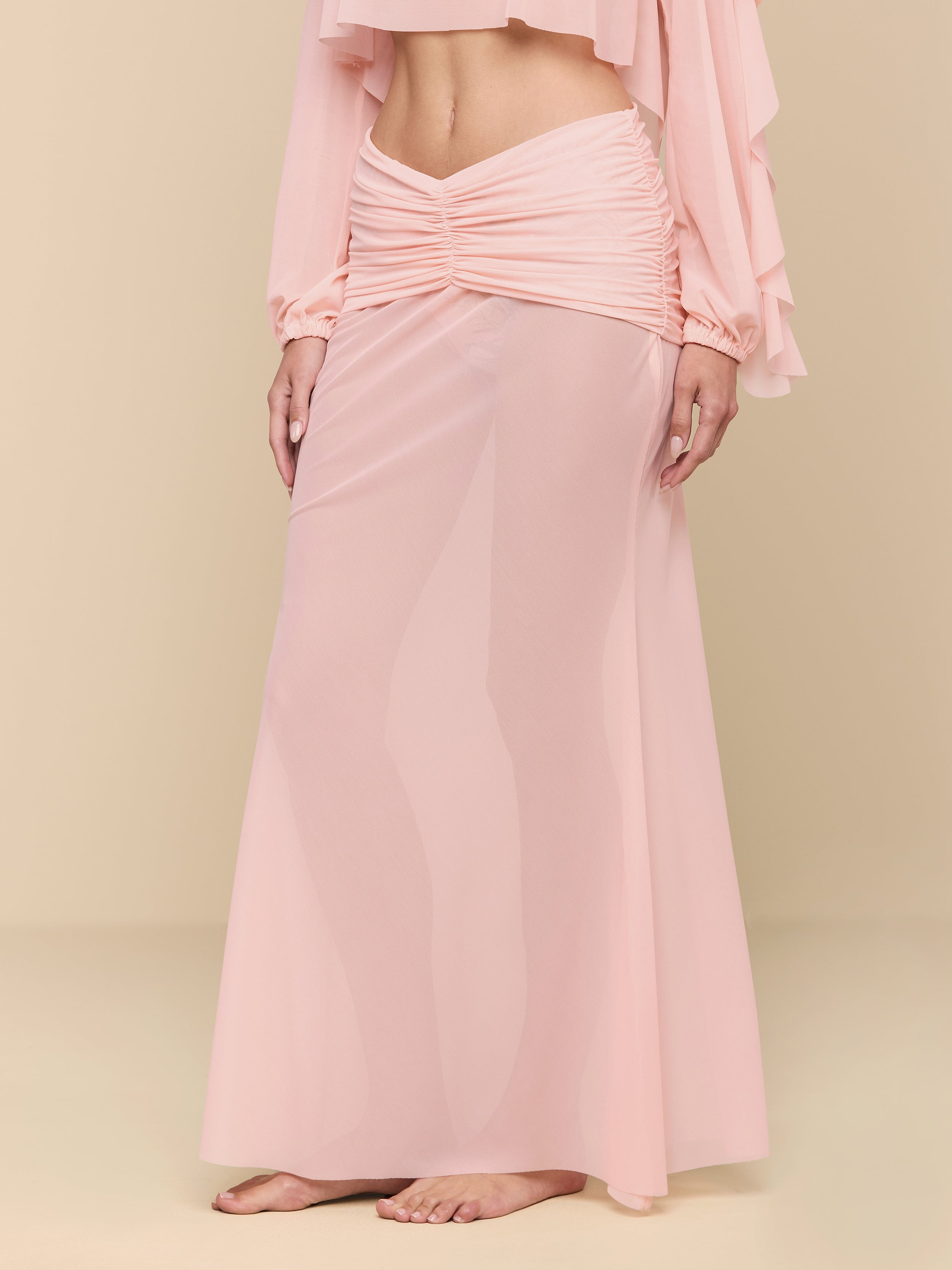A person stands barefoot wearing a long, sheer, light pink Mahe Mesh Cover-Up Skirt with ruching at the waist and a matching cropped top. The background is plain white.