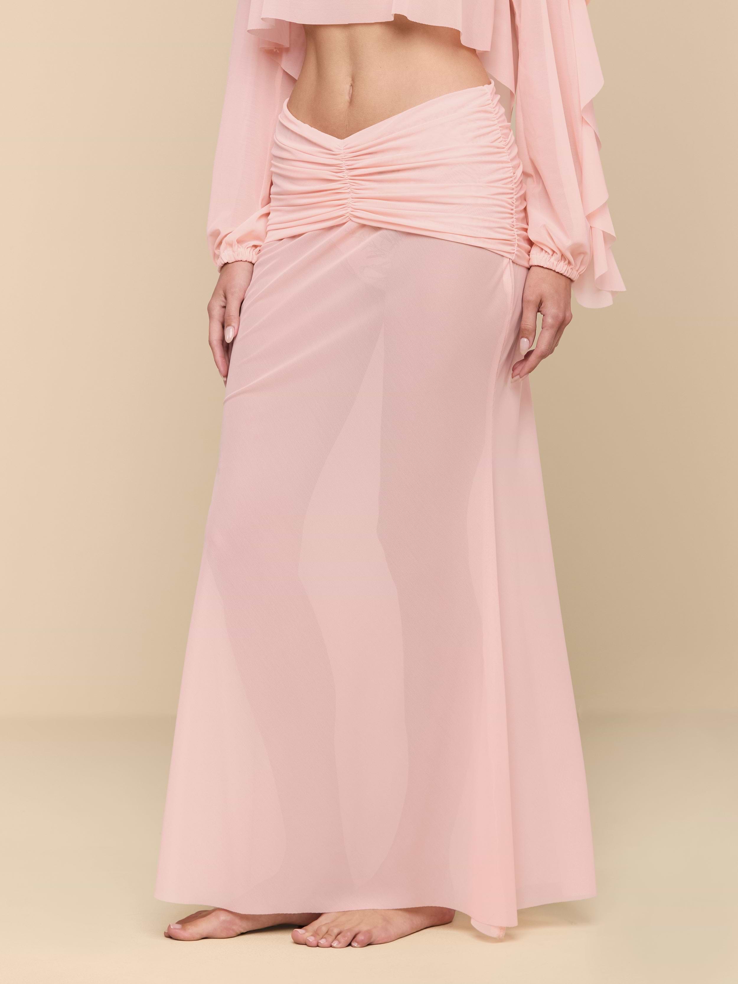 A person stands barefoot wearing a long, sheer, light pink Mahe Mesh Cover-Up Skirt with ruching at the waist and a matching cropped top. The background is plain white.