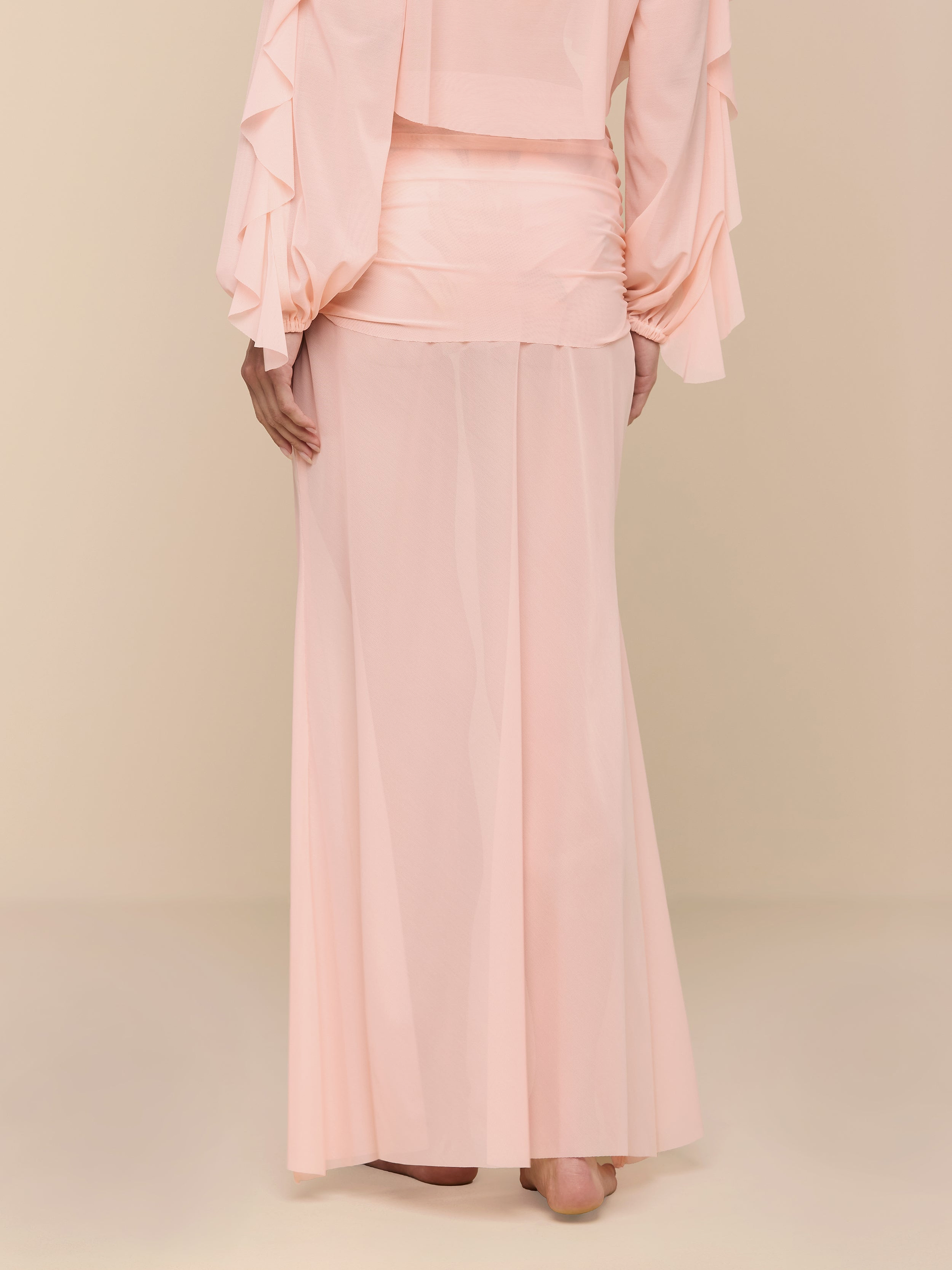 A person wearing a light pink, long-sleeved blouse and a matching pink Mahe Mesh Cover-Up Skirt stands barefoot against a plain white background, with their hands resting at their sides.