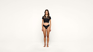 A woman stands facing forward against a plain white background, wearing a black crop top and black underwear, with her arms relaxed at her sides.