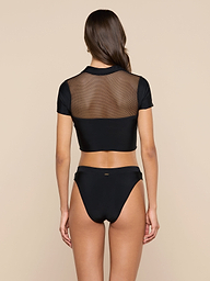 A woman stands with her back to the camera, showcasing the Anastasia Swim Top—a black short-sleeve mesh crop top—paired with black bikini bottoms for a look of beachside sophistication against a plain light background.