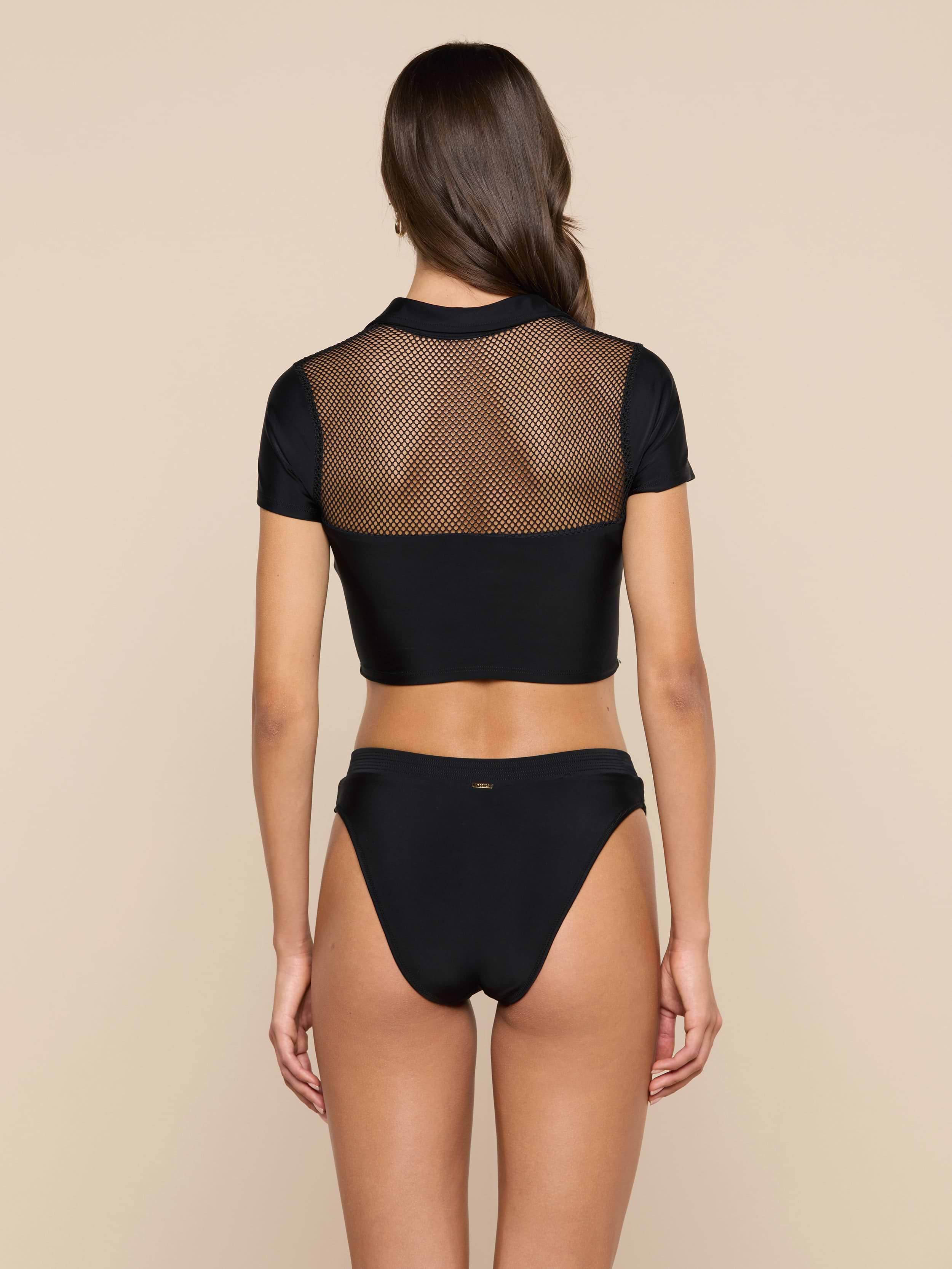 A woman stands with her back to the camera, showcasing the Anastasia Swim Top—a black short-sleeve mesh crop top—paired with black bikini bottoms for a look of beachside sophistication against a plain light background.