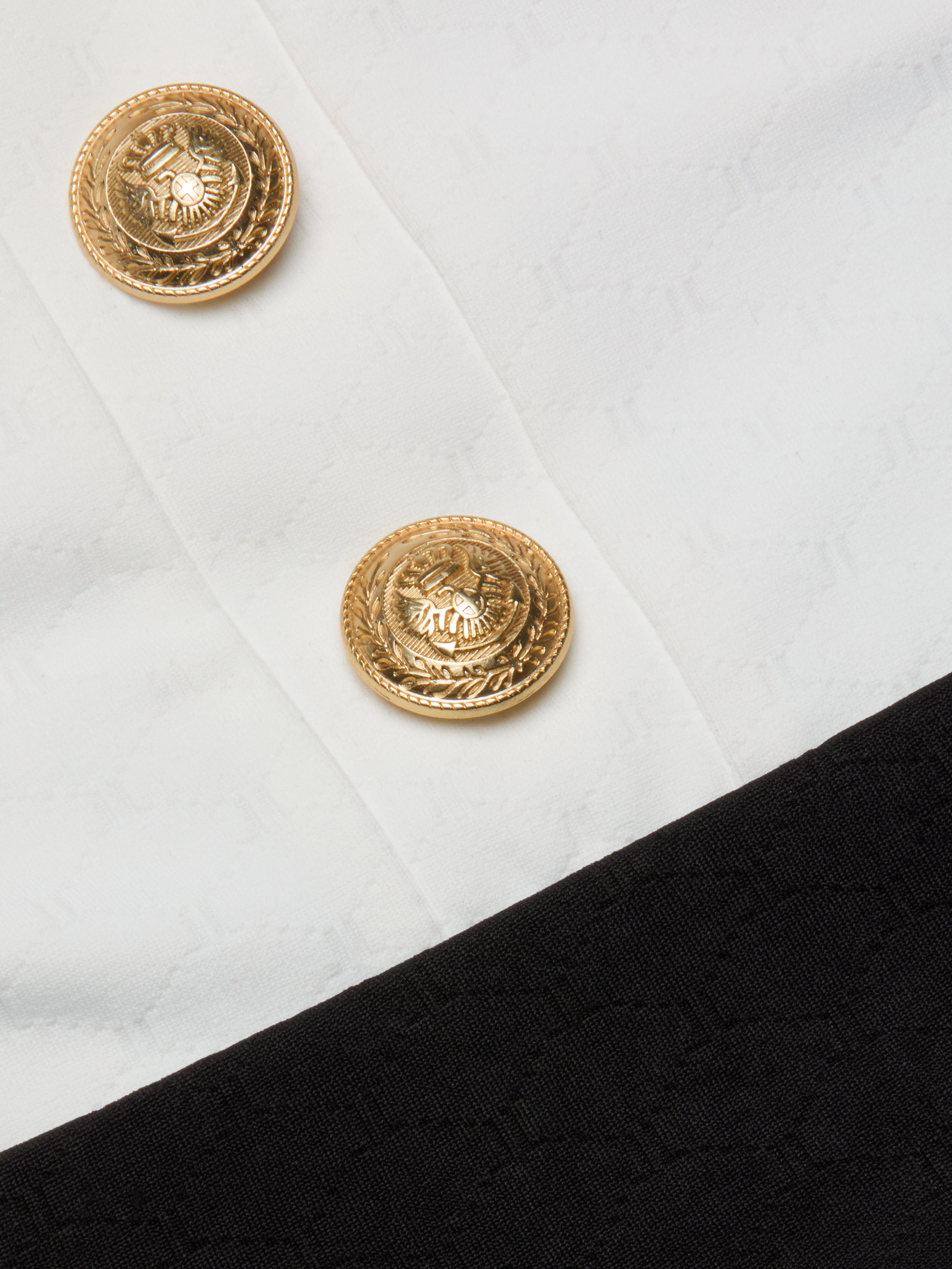 Close-up of two gold decorative buttons with an eagle emblem on the Mila One-Piece Swimsuit, set against textured white fabric with a black border at the bottom right corner, evoking a touch of French glamour.