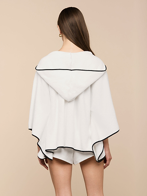 A woman with long brown hair stands facing away against a beige background, wearing the Demeter Cover-Up Poncho—a white terrycloth hooded poncho with black trim—paired with white shorts.