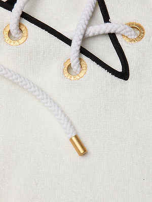 Close-up of a white braided drawstring with gold metal tip, threaded through gold-rimmed eyelets on soft terrycloth fabric. A black cord from the Demeter Cover-Up Poncho crosses over the white string.
