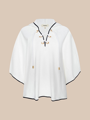 The Demeter Cover-Up Poncho is a white hooded spa poncho with a lace-up front, gold eyelets and gold-tipped drawstrings. Made from soft terrycloth, it features black trim on the hem and neckline, shown against a beige background.