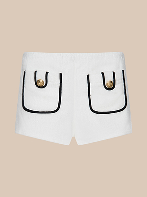 The Amiya Cover-Up Short features white terrycloth, two front pockets with black trim, gold button closures, and is perfect for chic poolside lounging. Shown here against a beige background.