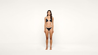 A woman with long brown hair stands barefoot against a plain white background, wearing a black and white bikini.