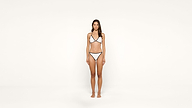 A woman stands barefoot against a plain white background, wearing a white bikini with black trim. She faces forward with a neutral expression and her arms relaxed at her sides.
