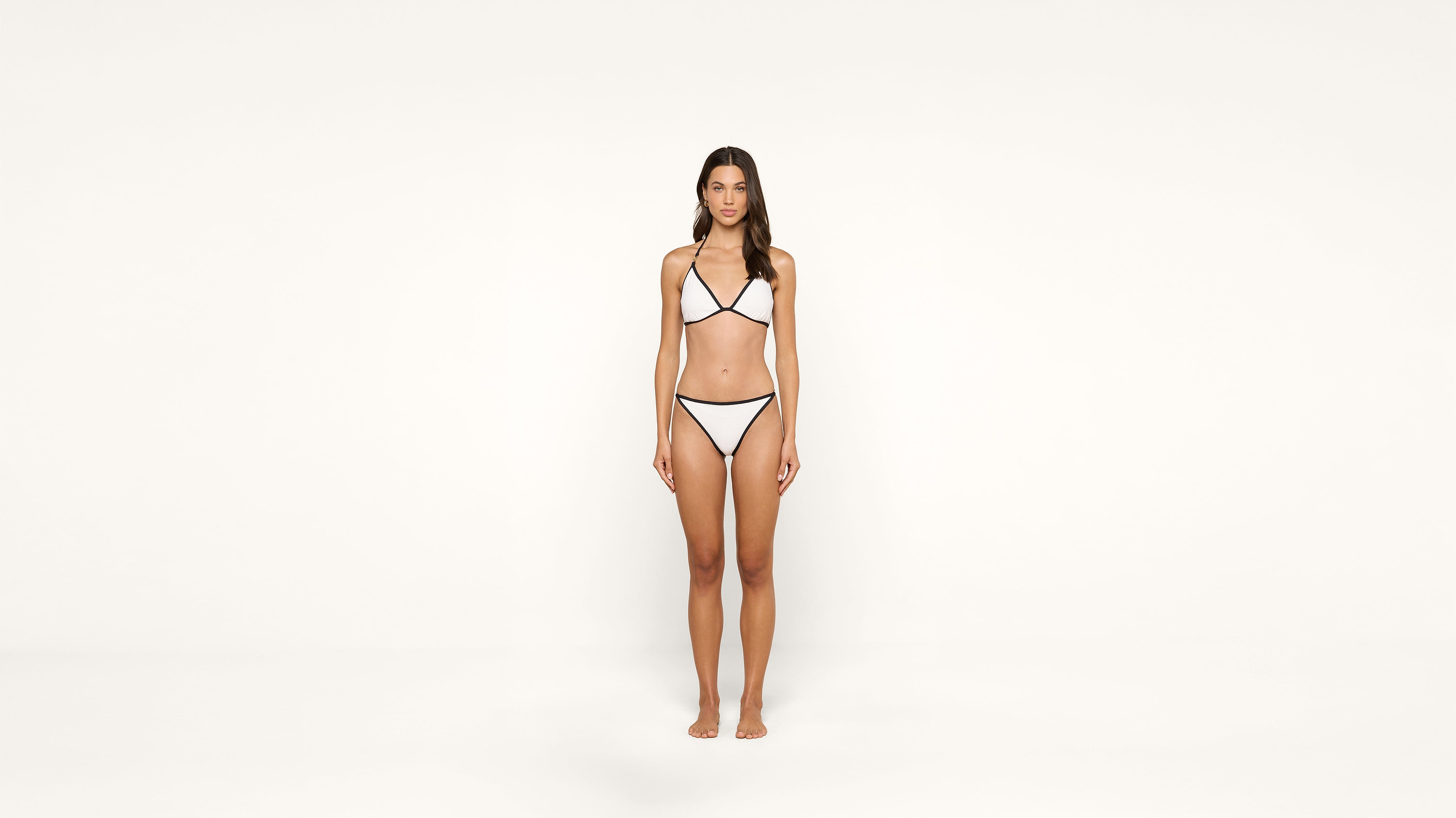 A woman stands barefoot against a plain white background, wearing a white bikini with black trim. She faces forward with a neutral expression and her arms relaxed at her sides.
