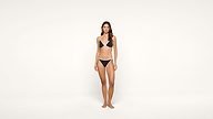 A woman stands barefoot against a plain white background, wearing a black bikini with white trim.